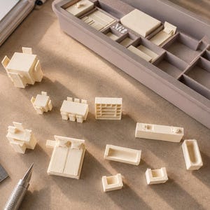 May include: A collection of off-white 1:100 scale miniature furniture pieces including tables, chairs, beds, and shelving units. The set is organized in a gray compartmentalized storage tray, with a metal ruler showing metric measurements.