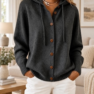 May include: A dark charcoal gray knit hooded cardigan featuring a button-down front with brown circular buttons, long sleeves with ribbed cuffs, a matching ribbed hem, and a drawstring hood. The garment is paired with plain white trousers.