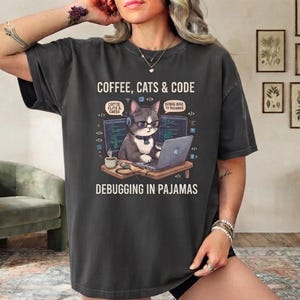 May include: A charcoal gray t-shirt featuring a graphic of a cat wearing glasses and headphones while working on a laptop. The text reads COFFEE, CATS & CODE and DEBUGGING IN PAJAMAS, with speech bubbles containing COFFEE, CATS & CODE and DEBUGGING IN PAJAMAS.