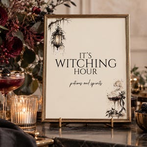 May include: A rectangular sign in a gold frame featuring the black text IT'S WITCHING HOUR and potions and spirits. The sign includes a black ink illustration of a hanging lantern in the top left corner and a cocktail glass in the bottom right.