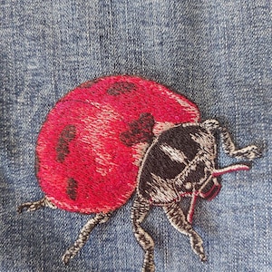 Op de afbeelding: Een blauwe spijkerstof met een gedetailleerd geborduurd lieveheersbeestje. Het lieveheersbeestje heeft felrode vleugels met zwarte stippen en een zwarte kop en borststuk met witte accenten. Het insect is afgebeeld met zichtbare pootjes.