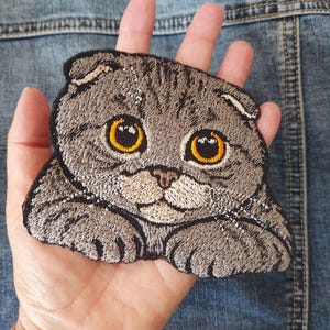 Peut inclure: Un écusson brodé représentant un chat Scottish Fold gris avec de grands yeux ronds ambrés et des oreilles repliées. Le chat est représenté avec ses pattes avant posées vers l'avant, avec des détails en fils gris, blancs et noirs.