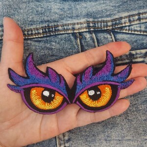 Könnte beinhalten: Ein gestickter Aufnäher mit zwei stilisierten Augen, leuchtend orangefarbenen Iris, schwarzen Pupillen mit weißen Lichtpunkten sowie violetten und blauen, federartigen Augenlidern. Das Design ist symmetrisch und hat oben spitze Kanten.