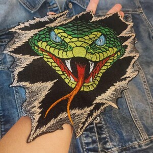 May include: An embroidered patch featuring a green snake head with blue eyes, white fangs, and a red and orange forked tongue. The snake appears to be emerging from a black background with jagged, ripped edges in shades of grey and white thread.