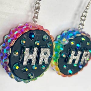 May include: A pair of dangle earrings featuring circular black bases with the letters HR in silver rhinestones. The edges are decorated with iridescent, multi-colored crystals. Each earring hangs from a silver-toned metal chain and hook.