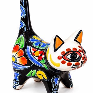 May include: A ceramic cat figurine with a glossy black body and a white face. The body features vibrant hand-painted floral patterns in orange, yellow, blue, and green. The white face has orange-lined ears and red dot accents around the eyes.