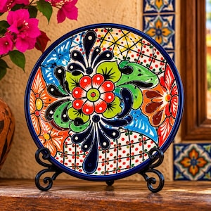 May include: A round ceramic plate featuring a vibrant, hand-painted floral design with a central red flower, green leaves, and orange accents. The plate has a dark blue rim and rests on a black metal display stand with scrolled feet.