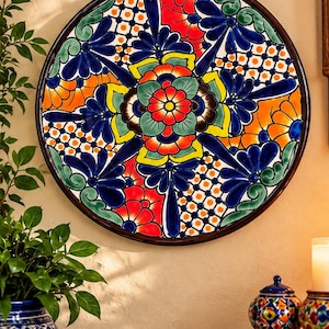 May include: A circular ceramic wall plate featuring a vibrant, hand-painted floral pattern in shades of red, orange, yellow, green, and deep blue. The design includes intricate geometric grid sections and a central multi-layered flower motif.