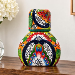 May include: A ceramic bedside carafe and matching tumbler set featuring a vibrant, hand-painted floral pattern in shades of orange, red, green, blue, and white. The carafe has a rounded base, and the tumbler sits inverted on top of the opening.