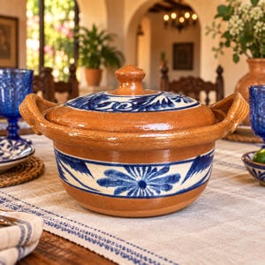 Puede incluir: Una cazuela redonda de terracota con tapa a juego, que presenta dos asas laterales y un patrón floral azul pintado a mano sobre una banda blanca alrededor del centro y la tapa. El acabado de arcilla natural es visible en el borde.