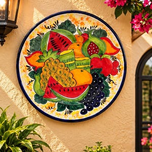 May include: A round ceramic wall plate featuring a vibrant hand-painted design of assorted fruits including watermelon slices, a pineapple, grapes, and oranges, set against a yellow and orange speckled background with a dark blue rim.
