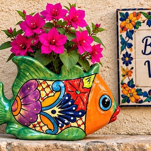 May include: A ceramic fish-shaped planter featuring a vibrant, multi-colored hand-painted pattern in green, orange, purple, and blue, containing a bright pink bougainvillea plant, positioned next to a decorative tile with the text Buenas Vibras.