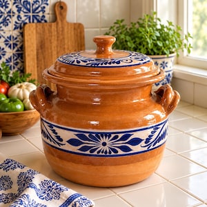 May include: A round, glazed terracotta cooking pot with two side handles and a matching lid. The pot features a wide, cream-coloured band around the centre decorated with intricate blue floral patterns, which are also repeated on the lid.