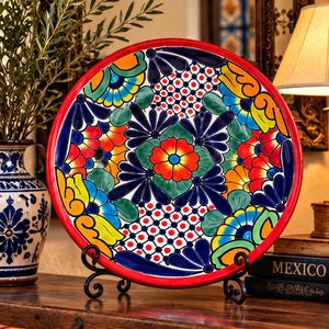 Peut inclure: Une assiette ronde en céramique ornée d'un motif floral et géométrique peint à la main dans des tons rouge, bleu, jaune, vert et orange. Le design présente une fleur centrale entourée de pétales complexes et un bord rouge sur un support noir.