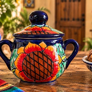 May include: A round ceramic sugar bowl with a lid, featuring a dark blue base color and a hand-painted sunflower design in vibrant shades of yellow, orange, and red, accented with green leaves and two curved side handles for easy gripping.
