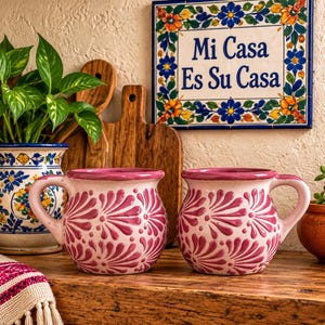 May include: Two round ceramic mugs featuring a light pink base with a dark pink hand-painted fan-like floral pattern. Each mug has a curved handle and a wide rim. Behind them sits a square tile with blue and yellow flowers and the text Mi Casa Es Su Casa.