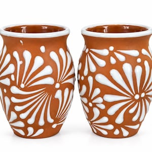 May include: Two terracotta-colored ceramic cups featuring a white painted floral pattern. Each cup has a rounded body, a wide opening with a white rim, and a smooth finish. The white design consists of radiating petal-like shapes.