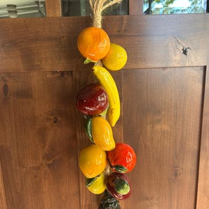 Könnte beinhalten: Ein hängendes Dekorationsobjekt bestehend aus einer Ansammlung glasierter Keramikfrüchte an einem dicken Juteseil. Das Arrangement umfasst eine leuchtende Orange, eine gelbe Zitrone, eine gelbe Banane, einen roten Apfel, eine gelbe Mango, eine rote Tomate und eine texturierte Ananas.