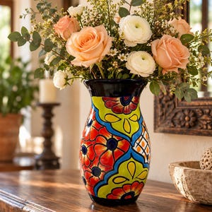 May include: A ceramic vase featuring a vibrant, hand-painted floral pattern in bright red, lime green, and deep blue. The vase has a flared rim and a glossy finish, filled with a bouquet of peach-colored roses and white ranunculus flowers.