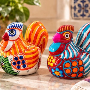 May include: Two ceramic rooster-shaped salt and pepper shakers featuring vibrant hand-painted designs. The left shaker has orange, yellow, and green patterns, while the right shaker displays red, blue, and white stripes with floral motifs.