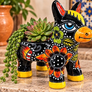 May include: A black ceramic donkey-shaped planter featuring vibrant hand-painted floral patterns in orange, yellow, and green, with white polka dots. The donkey has a bright orange snout, blue eyes, and yellow hooves, holding a green succulent plant.