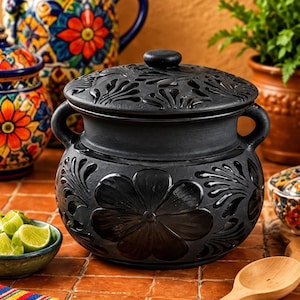 May include: A matte black ceramic pot with a matching lid, featuring two side handles and embossed floral patterns on the body and lid. The pot has a rounded shape and a smooth finish, showcasing intricate carved petal designs across its surface.