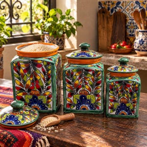 May include: Three square ceramic canisters with lids, featuring hand-painted green, yellow, red, and blue floral and leaf patterns. The canisters have orange rims and matching lids with knobs. A small wooden scoop sits nearby with grains.