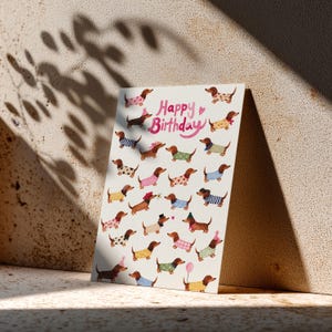 Könnte beinhalten: Eine rechteckige Grußkarte mit cremefarbenem Hintergrund, die den rosa Schriftzug Happy Birthday und zahlreiche Illustrationen von Dackeln in verschiedenen bunten Pullovern, Hüten und Accessoires auf der Kartenoberfläche zeigt.