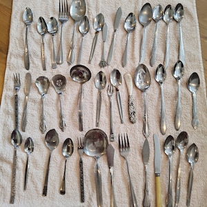 May include: A selection of vintage cutlery, including spoons, forks, and knives, displayed on a light-coloured cloth. The silverware features diverse handle designs and sizes, showcasing a range of styles from classic to ornate.