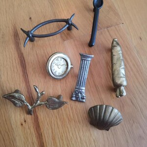 May include: Assortment of decorative cabinet knobs in various styles and finishes, including a clock face, a seashell, a column, a leaf design, a spoon, and a barbed wire design.