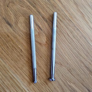 May include: Two silver metal tools with textured handles and different shaped ends. One has a flat end, and the other has a rectangular end. The tools are on a wooden surface.