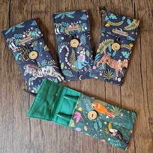 Könnte beinhalten: Stoff-Stifteetuis mit Dschungeltier-Mustern. Die Etuis zeigen Zebras, Tiger und Lemuren auf marineblauem Hintergrund. Ein Etui ist geöffnet und zeigt grüne Stifte. Jedes Etui hat einen Verschluss mit Holzknopf.