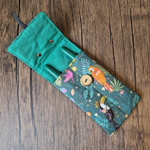 Puede incluir: Estuche para bol&iacute;grafos de tela verde con solapa asegurada por un bot&oacute;n de madera. El estuche presenta un colorido estampado de aves tropicales y flores. Dos bol&iacute;grafos verdes son visibles dentro del estuche abierto. Se adjunta un lazo el&aacute;stico negro.