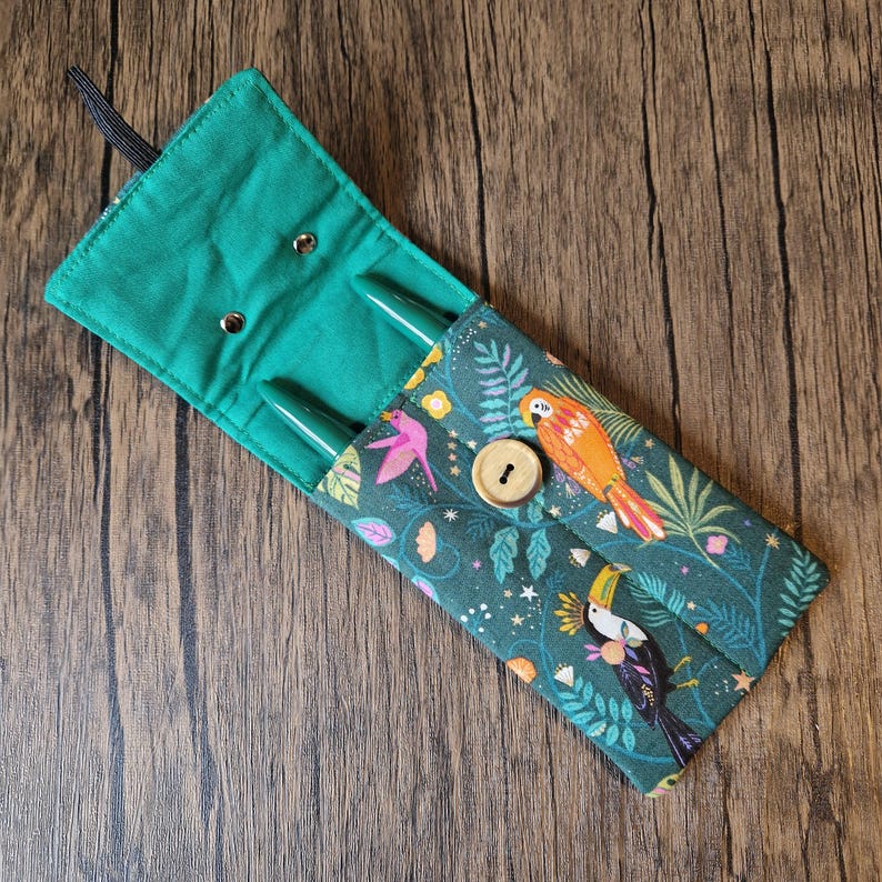 Puede incluir: Estuche para bol&iacute;grafos de tela verde con solapa asegurada por un bot&oacute;n de madera. El estuche presenta un colorido estampado de aves tropicales y flores. Dos bol&iacute;grafos verdes son visibles dentro del estuche abierto. Se adjunta un lazo el&aacute;stico negro.