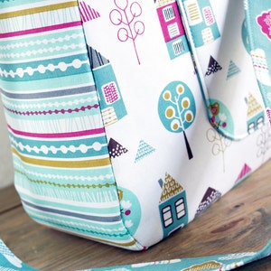May include: A turquoise tote bag with a white background and a colourful pattern of houses, trees, and geometric shapes. The bag has a turquoise strap with a cloud pattern.