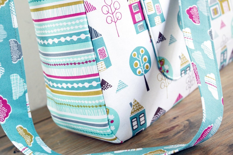 May include: A turquoise tote bag with a white background and a colourful pattern of houses, trees, and geometric shapes. The bag has a turquoise strap with a cloud pattern.