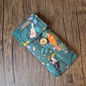 Puede incluir: Estuche de tela verde azulado con solapa, con un colorido estampado de aves tropicales. El estuche se asegura con un bot&oacute;n de madera y tiene una etiqueta met&aacute;lica que dice "SusieDDesigns". El dise&ntilde;o incluye loros, tucanes y elementos florales.