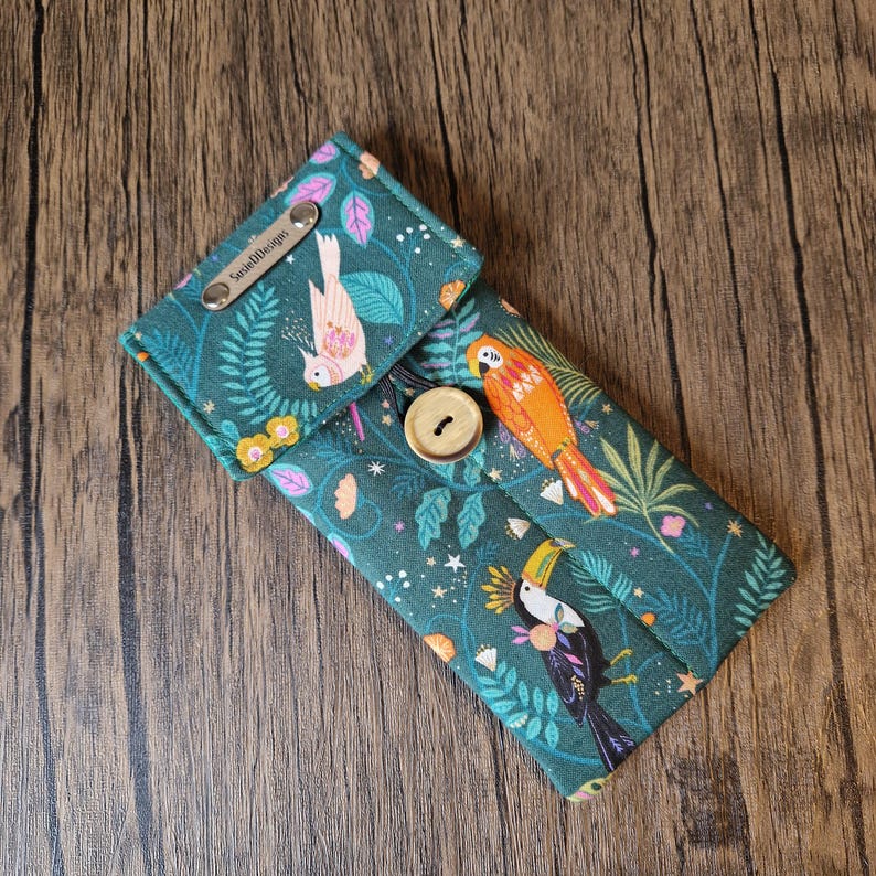 Puede incluir: Estuche de tela verde azulado con solapa, con un colorido estampado de aves tropicales. El estuche se asegura con un bot&oacute;n de madera y tiene una etiqueta met&aacute;lica que dice "SusieDDesigns". El dise&ntilde;o incluye loros, tucanes y elementos florales.