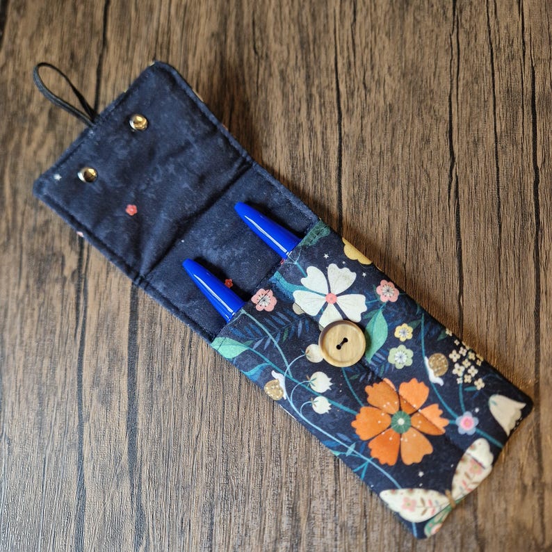 Puede incluir: Un estuche para bol&iacute;grafos de tela azul marino con un estampado floral, con flores naranjas y blancas, hojas verdes y un cierre de bot&oacute;n de madera. Se ven dos bol&iacute;grafos azules en el interior. El estuche tiene un lazo negro para colgar.
