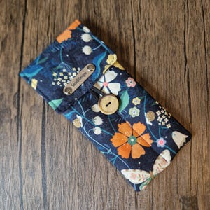 Puede incluir: Un estuche para gafas azul marino con estampado floral con flores naranjas, blancas y amarillas. Tiene un cierre de bot&oacute;n de madera y una peque&ntilde;a etiqueta met&aacute;lica con "SusieDDesigns".