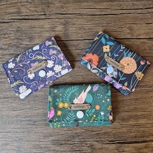 Può includere: Tre porta carte in tessuto con motivi floreali e di uccelli. Ogni portafoglio è dotato di una chiusura a scatto e di una piccola etichetta in pelle con la scritta "SusieDDesigns". I portafogli hanno uno sfondo blu scuro, verde scuro e nero.