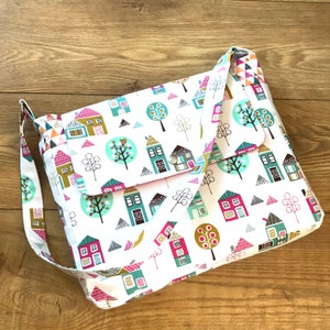 Könnte beinhalten: Weiße Umhängetasche mit einem farbenfrohen Muster aus Häusern und Bäumen. Die Tasche hat einen Klappverschluss mit Knopf und einen langen Riemen. Das Design umfasst rosa, türkis, braune und blaue Häuser und Bäume auf weißem Grund.