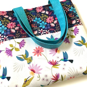 May include: A white tote bag with a floral pattern and hummingbirds. The bag has teal straps and a dark blue floral lining.
