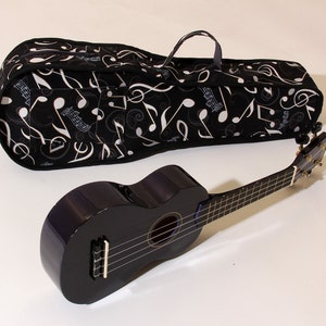 Könnte beinhalten: Eine schwarze Ukulele mit einem dunklen Holzgriffbrett und einer schwarzen und weißen Musiknoten-Muster-Gitarrentasche.