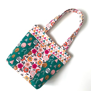 Könnte beinhalten: Eine kleine grüne, rosa, weiße und orangefarbene Tragetasche mit einem Blumen- und Baummuster. Die Tasche hat zwei Stoffgriffe und einen Druckknopfverschluss.