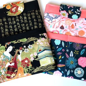 May include: Two fabric tablet sleeves with different designs. The first sleeve is black with gold accents and features a geisha playing a musical instrument. The second sleeve is pink and blue with a floral pattern.