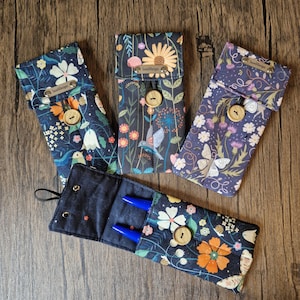 Puede incluir: Cuatro estuches de tela floral con cierres de bot&oacute;n de madera. Los estuches presentan coloridos estampados de flores y p&aacute;jaros sobre un fondo oscuro. Un estuche est&aacute; abierto, revelando dos bol&iacute;grafos azules. Cada estuche tiene una peque&ntilde;a etiqueta que dice "SueDesign".