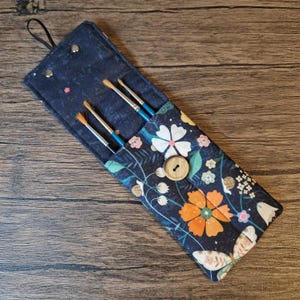 Puede incluir: Un estuche enrollable de tela azul marino con un estampado floral, que contiene cuatro pinceles. El rollo presenta flores blancas, naranjas y rosas, un cierre de bot&oacute;n de madera y un lazo negro para colgar. Los pinceles tienen mangos azules.