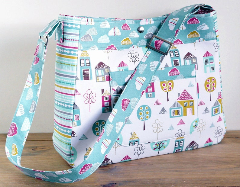 May include: A turquoise and white fabric tote bag with a long adjustable strap. The bag features a repeating pattern of houses, trees, and clouds. The bag has a flap with a snap closure.