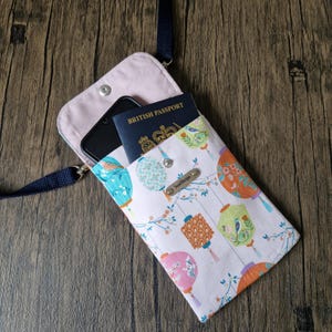 Peut inclure: Une housse de téléphone à bandoulière en tissu blanc avec un motif de lanternes colorées. L'étui est ouvert, révélant un smartphone noir et un passeport britannique. Une sangle bleu marine est attachée. Fermeture à bouton-pression.